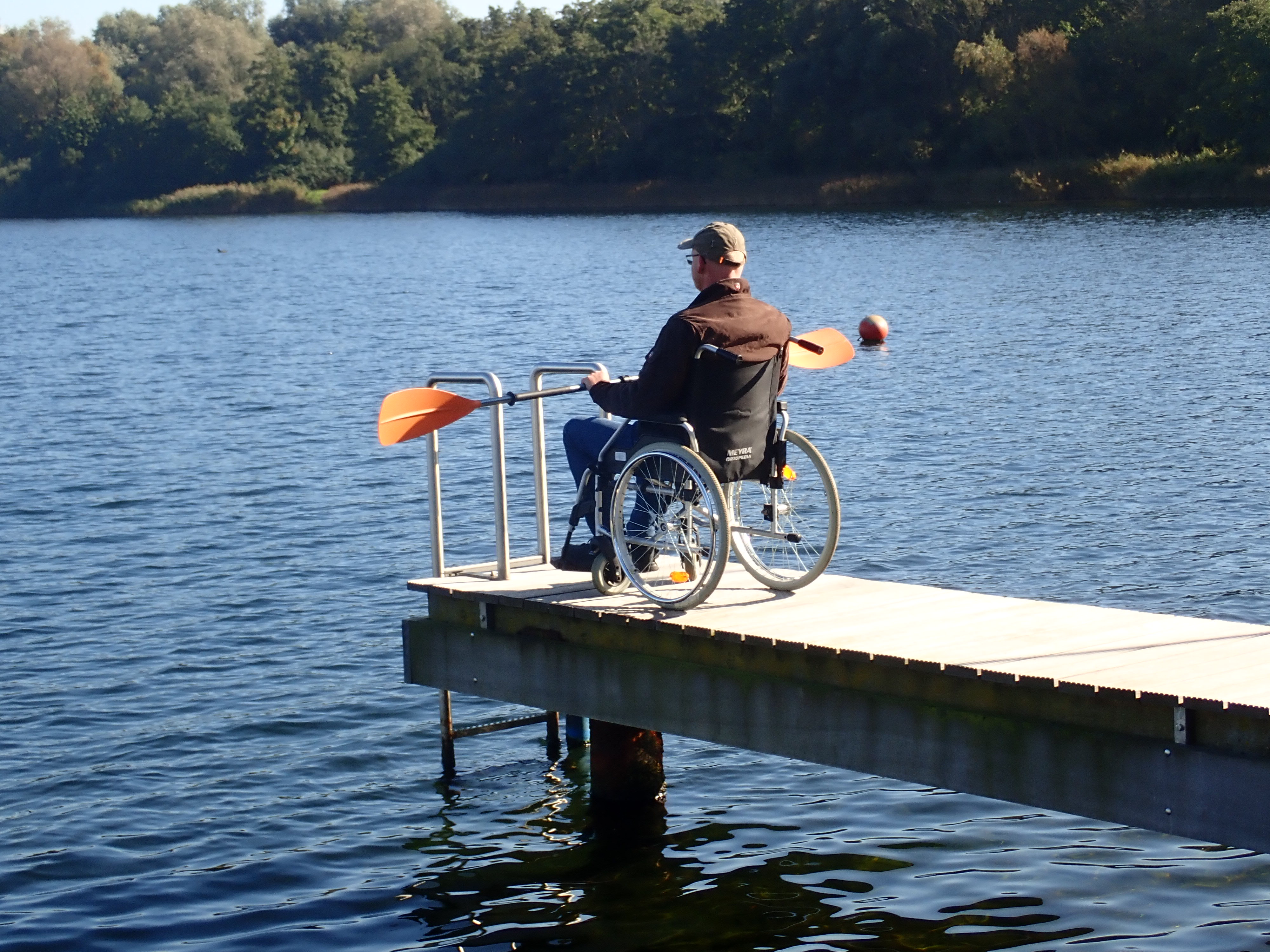 Ein Mann im Rollstuhl sitzt auf einem Steg am Wasser und hält ein Doppelpaddel vor sich