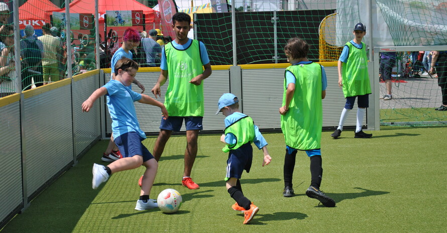 sechs Kinder und Jugendliche, vier davon in hellgrünen Leibchen, spielen Fußball auf einem von Banden begrenzten Spielfeld