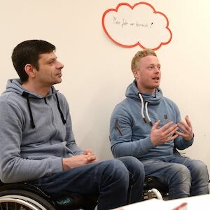 Zwei Projektteilnehmer sitzen vor einer Wand an der eine Gedankenwolke hängt. Der rechte Mann spricht, während der Linke interessiert zuhört. 