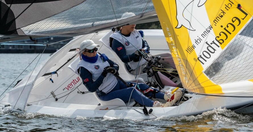zwei Personen mit norwegischer Flagge auf dem Trikot in einem schiefliegenden Segelboot mit gelbem Segel, auf dem ein Delfin abgebildet ist. 
