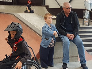 eine kleinwüchsige Frau und ein Mann inmitten eines Skateparks hinter einem Kind mit coolem Helm und Rollstuhl im Vordergrund