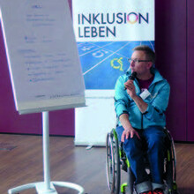 Thorsten Ely sitzt im Rollstuhl neben einem Flipchart-Ständer und hält einen Vortrag mit Mikrofon in der Hand. 
