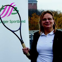 Ewa Astapczyk hält einen Tennisschläger und Ball hoch. Im Hintergrund sieht man auf einer Wand das Logo des Hamburger Sportbundes. 