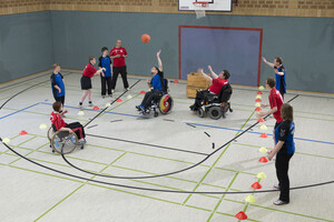 Rollstuhlfahrer*innen und Fußgänger in blauen und roten Trikots spielen unter dem Korb Basketball 