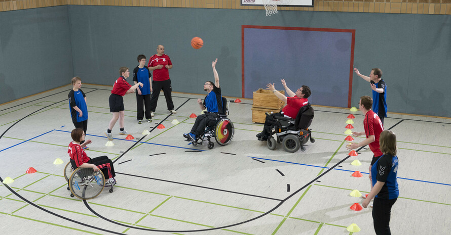 Rollstuhlfahrer*innen und Fußgänger in blauen und roten Trikots spielen unter dem Korb Basketball 
