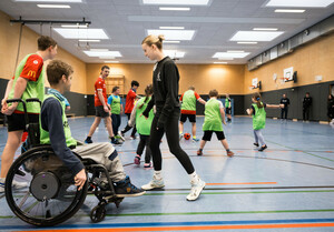 eine Gruppe von Kindern und Jugendlichen spielt in einer Sporthalle Fußball im Vordergrund zwei Personen und ein Rollstuhlfahrern